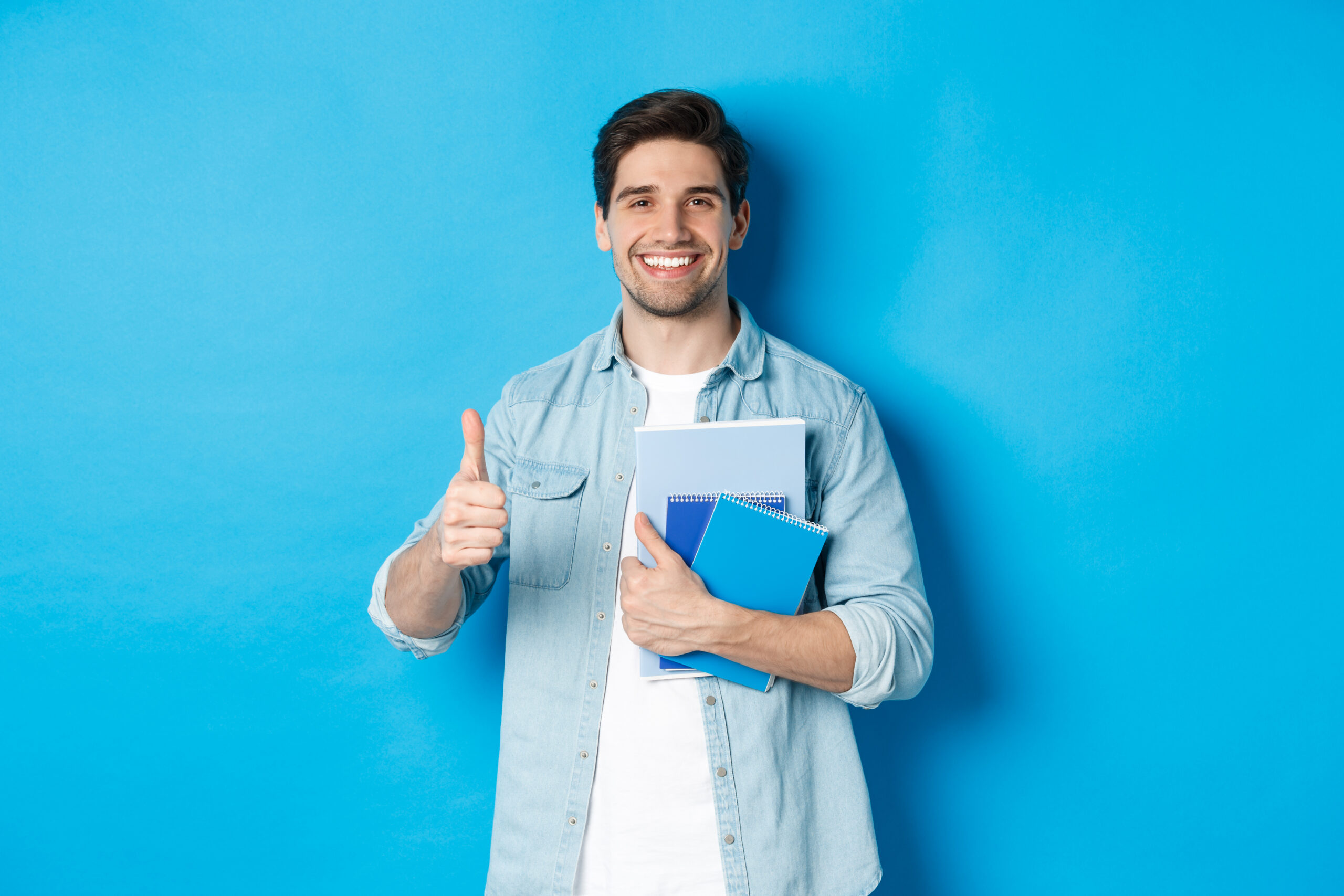 Young man student with notebooks, showing thumb up in approval, smiling satisfied, blue studio background.